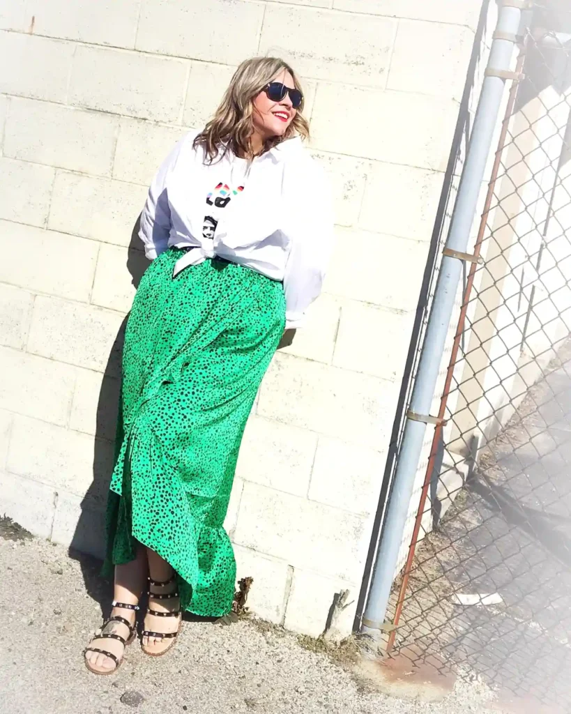 Woman in a white top and green skirt leaning on a wall, wearing sunglasses, smiling in the sunlight.