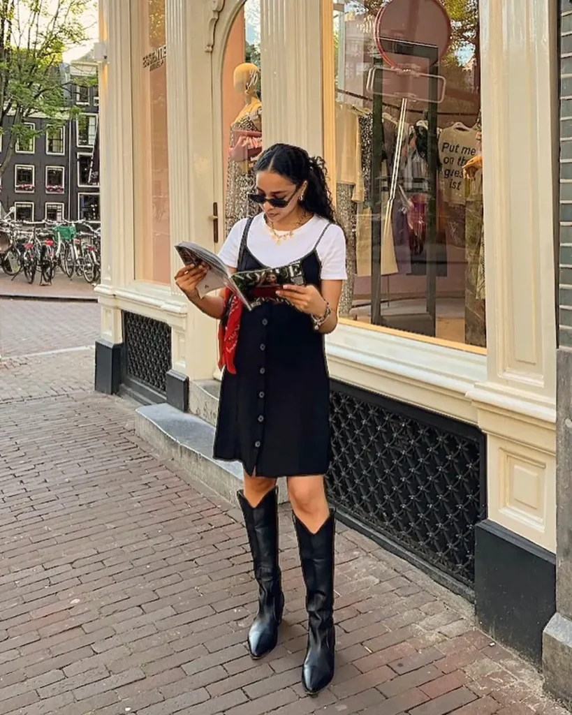 Stylish woman reading a magazine in front of a boutique, wearing a black dress and boots on a city street.