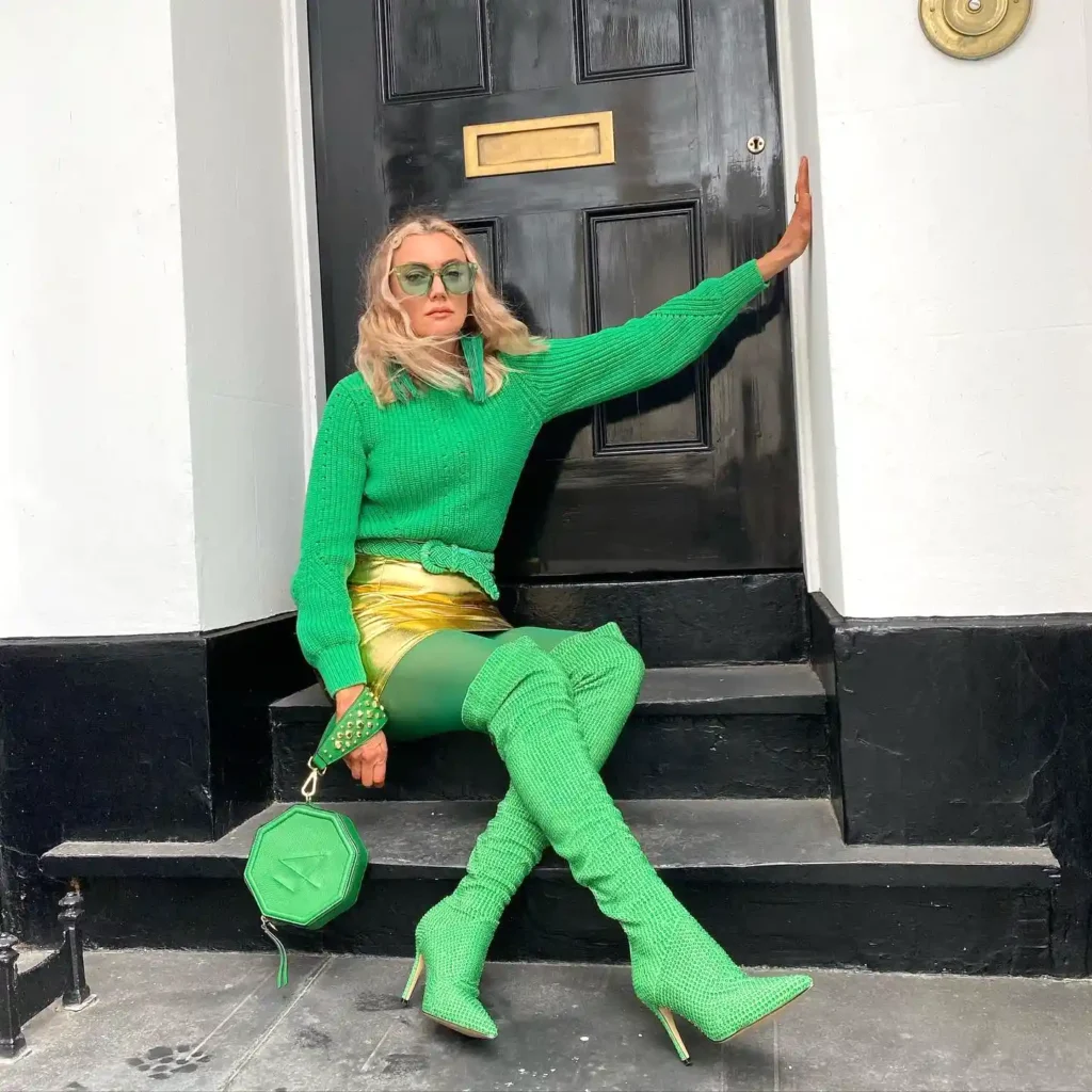 Fashionable woman in vibrant green outfit and boots on steps, holding geometric bag in front of a black door.