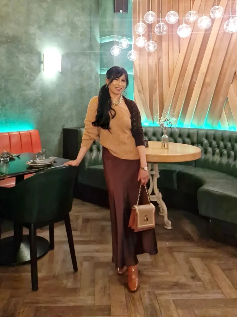 Stylish woman in a modern restaurant, wearing a brown outfit and holding a handbag, with elegant decor in the background.
