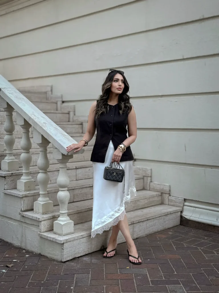 Woman standing by stairs in stylish black top and white skirt, holding a quilted purse.