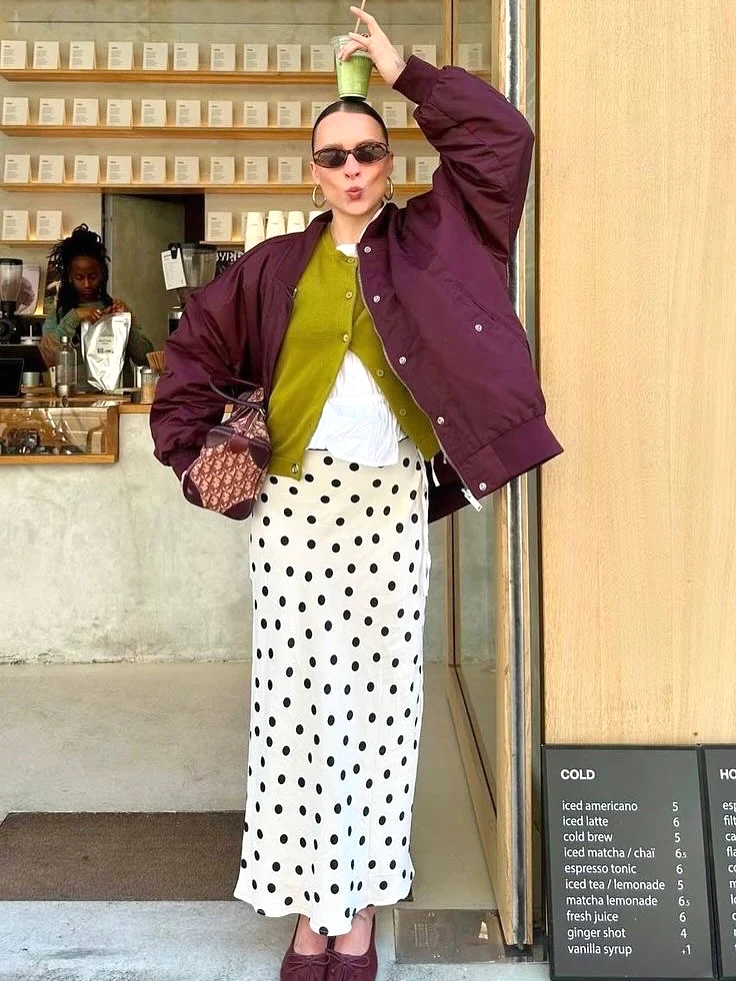 Fashionable woman poses playfully with smoothie, wearing a purple jacket and polka dot skirt outside a café.