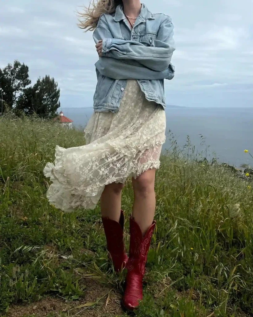 Person in lace dress, denim jacket, and red boots standing on grassy cliff with ocean view in the background.