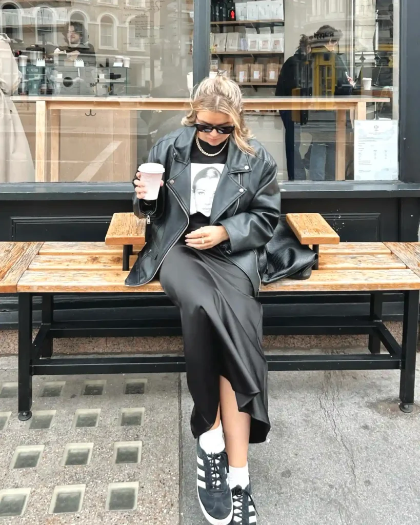 Woman in leather jacket and sneakers enjoys coffee on a bench outside a cafe.