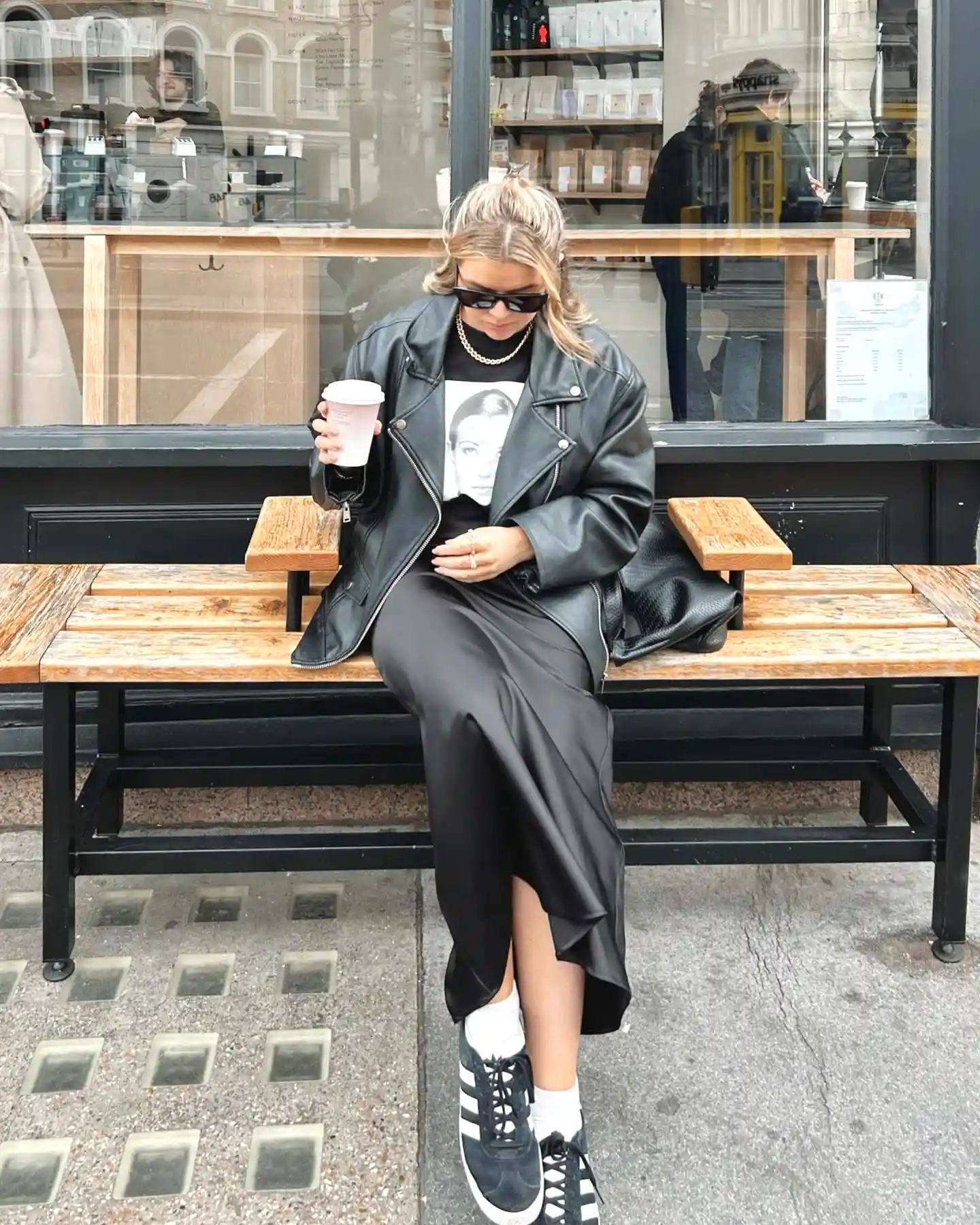 Woman in leather jacket and sneakers enjoys coffee on a bench outside a cafe.