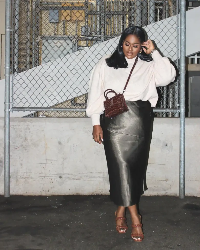 Fashionable woman in cream sweater and black skirt with a small brown purse, standing by a chain-link fence in urban setting.