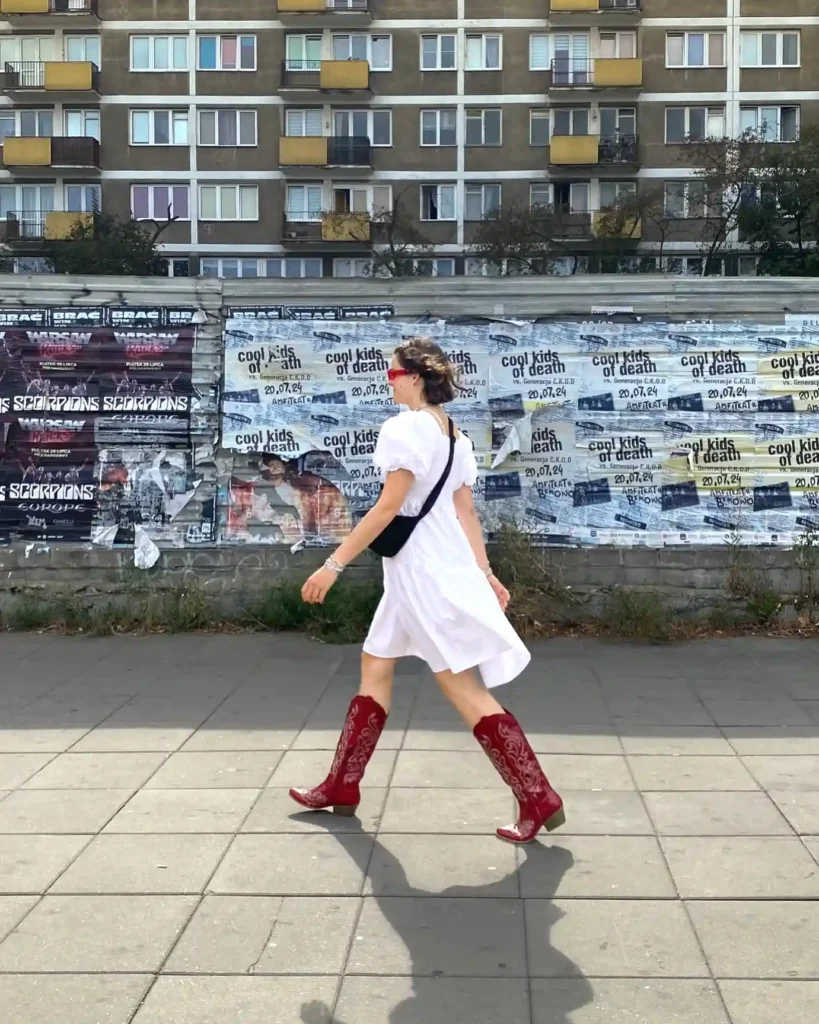 Woman in white dress and red boots walking past urban posters on street.