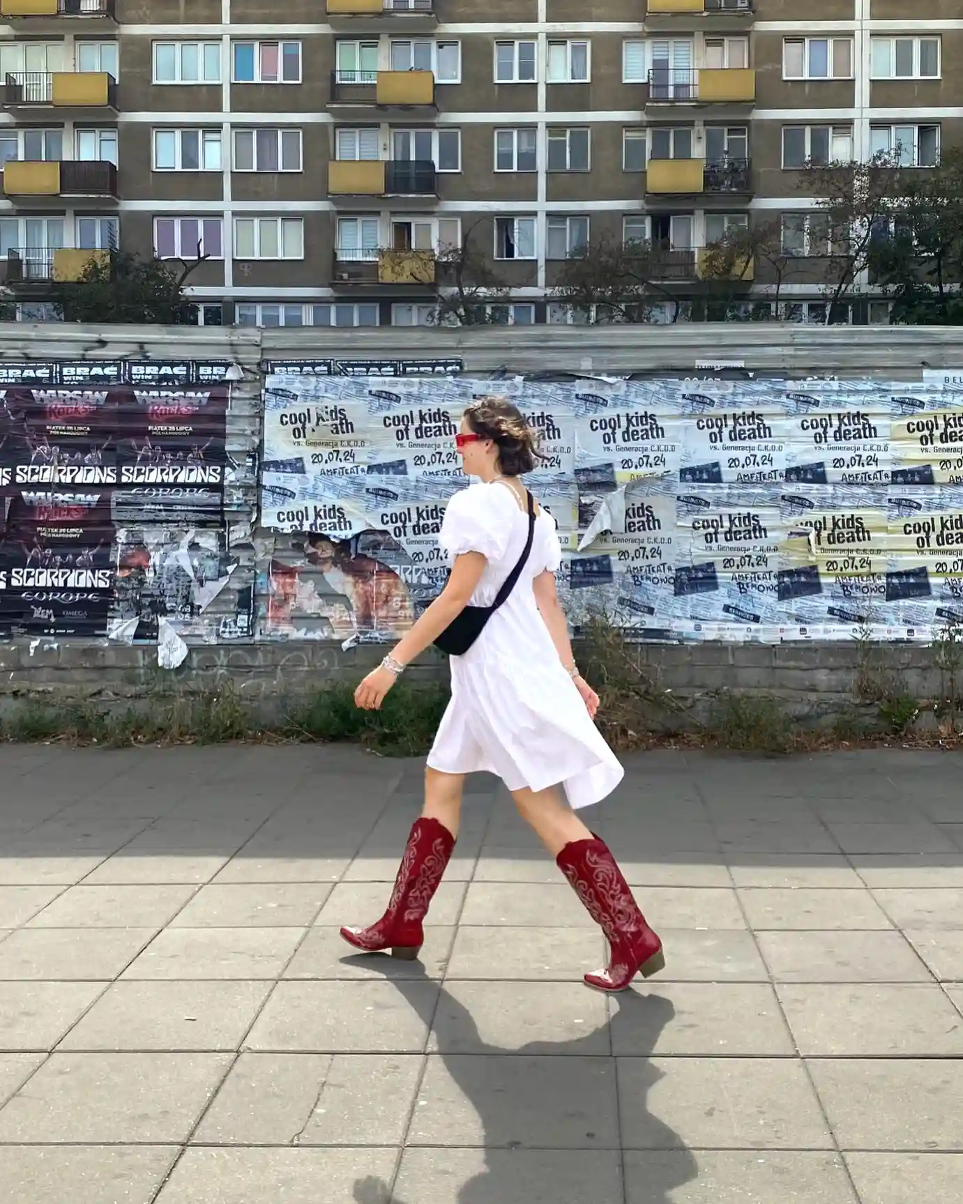 Woman in white dress and red boots walking past urban posters on street.