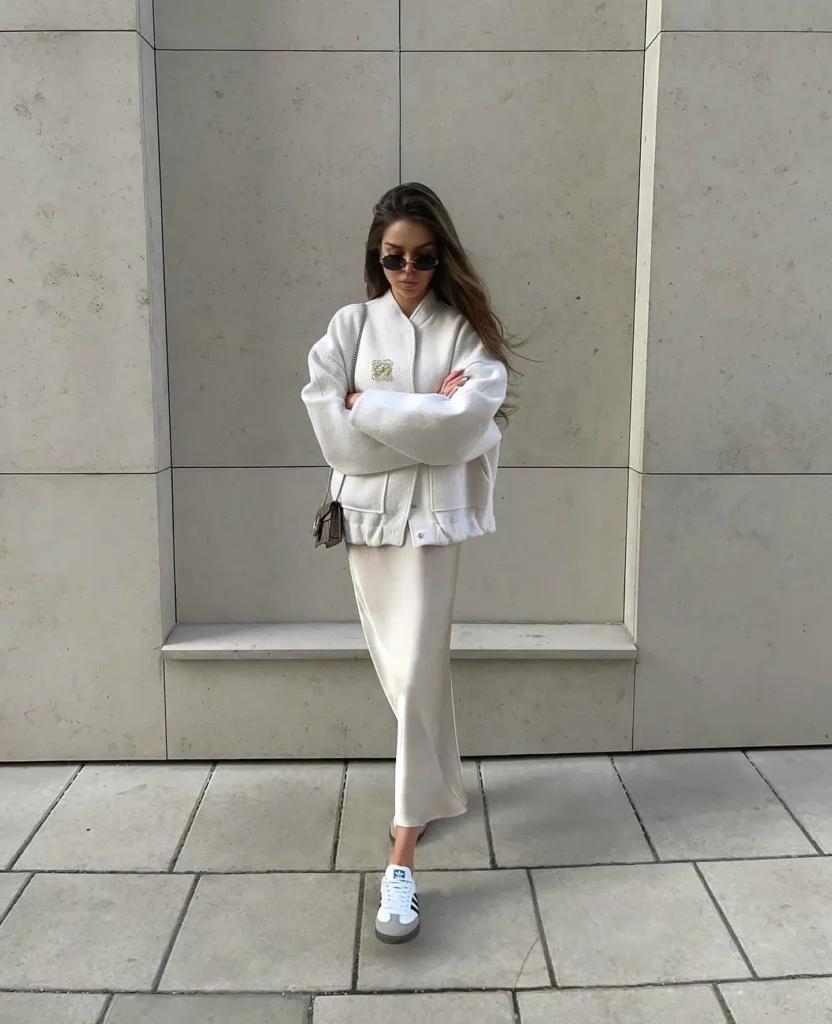 Stylish woman in white jacket, sunglasses, and sneakers posing against a modern beige wall.
