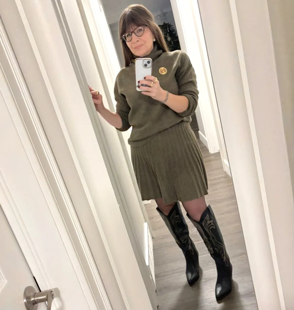 Person in mirror wearing a green sweater, pleated skirt, and black boots, taking a selfie in a hallway.