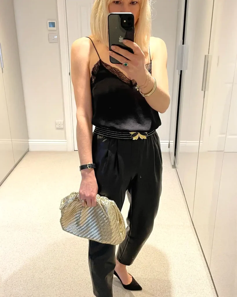 Woman in black outfit taking a selfie, accessorized with a gold clutch and jewelry in a mirror setting.