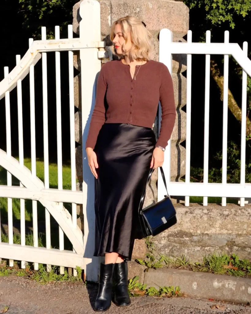 Woman in autumn fashion, wearing a brown top and black silk skirt, standing by a white fence on a sunny day.