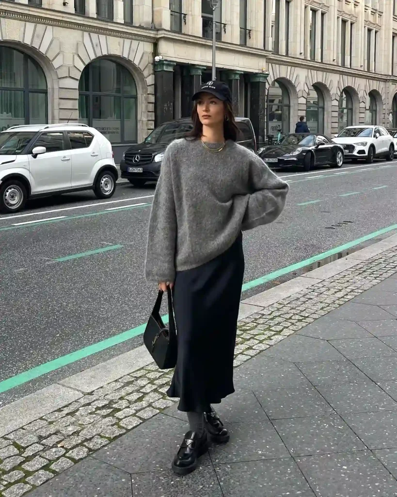 Woman in gray sweater and black skirt walking on urban street, holding black handbag, surrounded by parked cars.