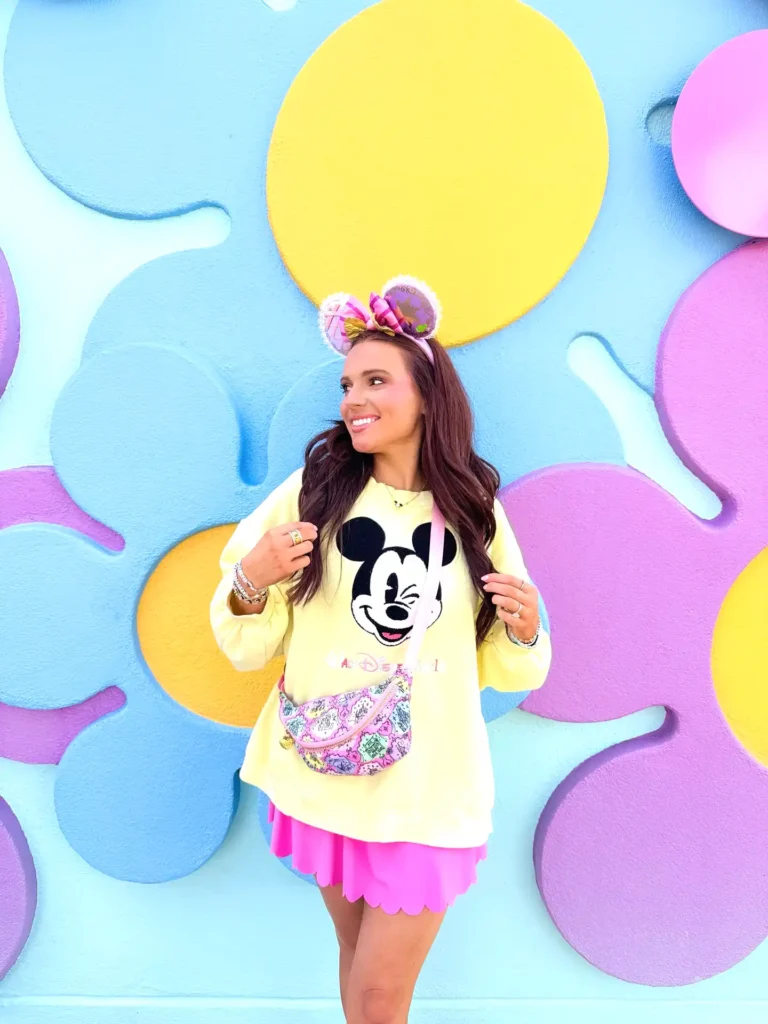 Woman in Mickey sweater and colorful outfit smiling against a vibrant pastel background.