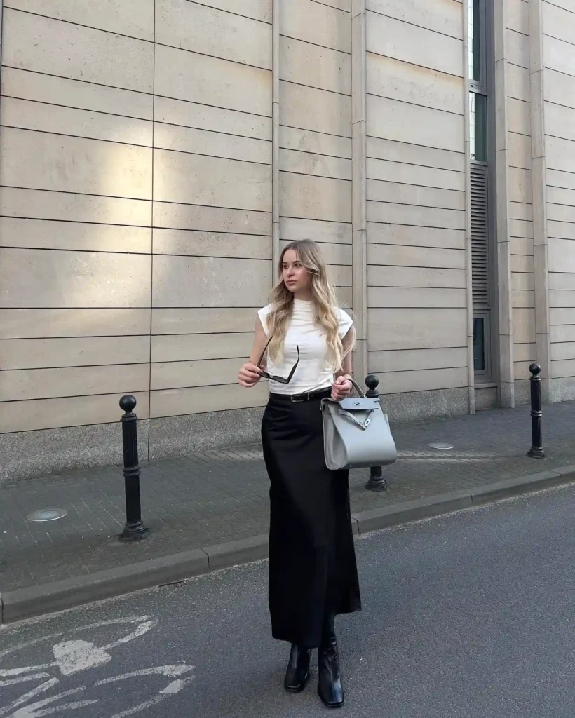Woman in stylish black skirt and white top holds gray handbag and sunglasses, walking by modern building.