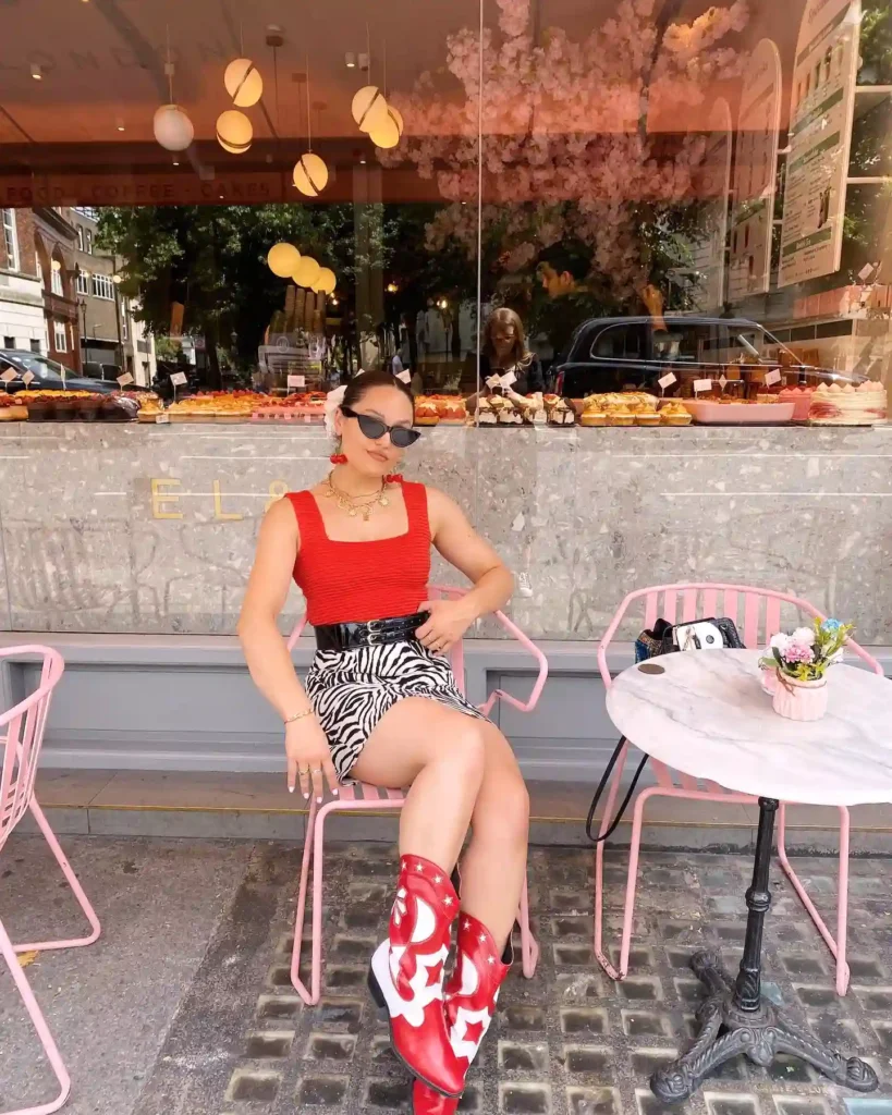 Stylish woman in red top and patterned skirt sits at outdoor café table with pink decor and floral centerpiece.