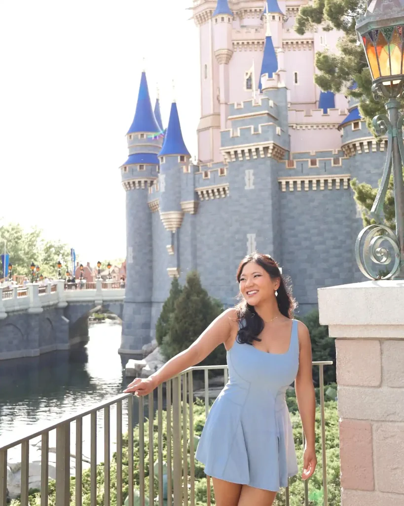 Person in blue dress smiling near a fairytale castle with blue spires, sunny day.