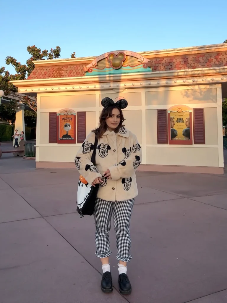 Person wearing Mickey Mouse ears and a patterned outfit stands in front of theme park entrance decorated for Halloween.
