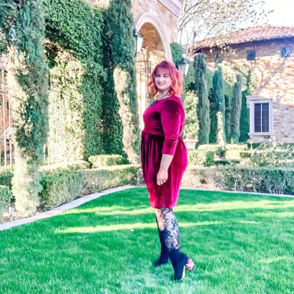 Woman in a red dress poses in a lush garden, near an ivy-covered stone building.