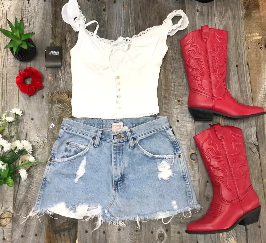 White lace top, denim skirt, red boots flat lay on wooden background with flowers and accessories. Rustic chic style.