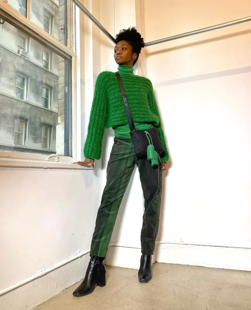 Fashionable person in green sweater, dark pants, and black boots poses by a window with city view, holding a stylish bag.