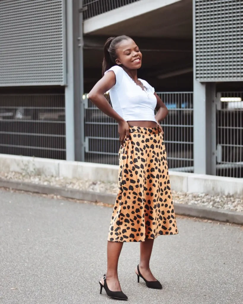Smiling woman in leopard print skirt and white top poses confidently outdoors near modern building.