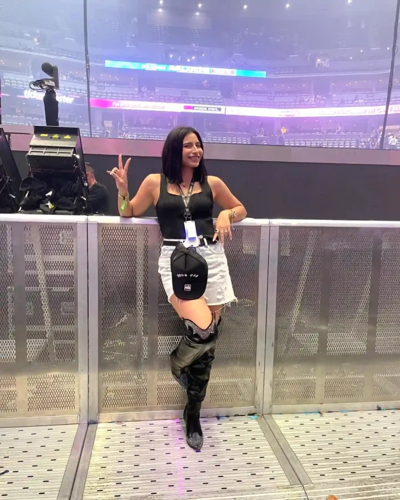 Woman posing by arena barrier, wearing black top, white skirt, and boots, flashing peace sign at concert.