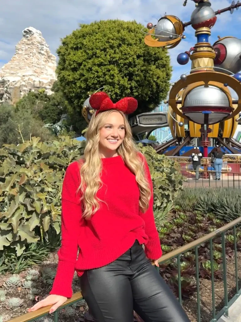 Woman in red sweater and ears, smiling at a theme park with rides and a mountain backdrop on a sunny day.