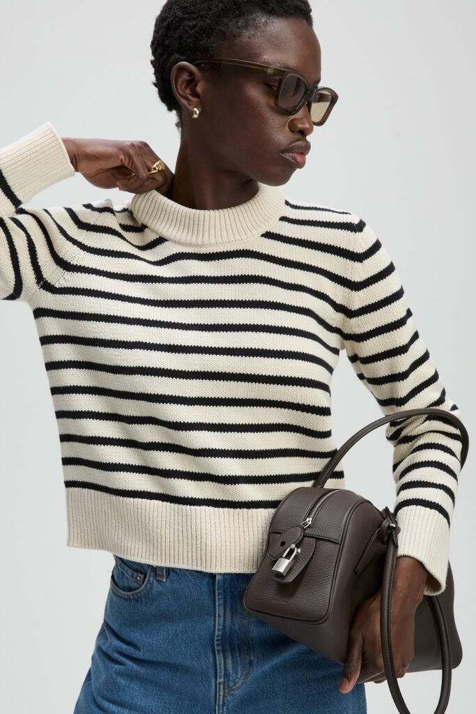 Model in striped sweater and denim skirt, holding a brown handbag, showcasing a stylish urban outfit.