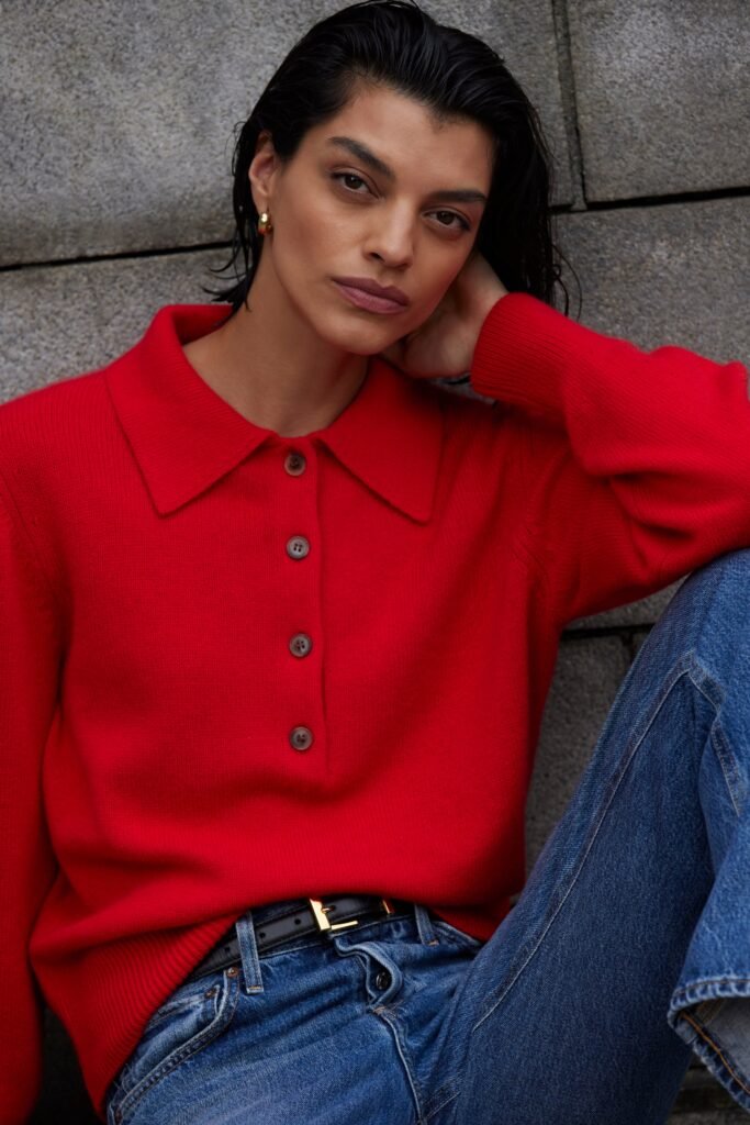 A model in a red sweater poses against a stone wall, showcasing a chic casual look with denim jeans.