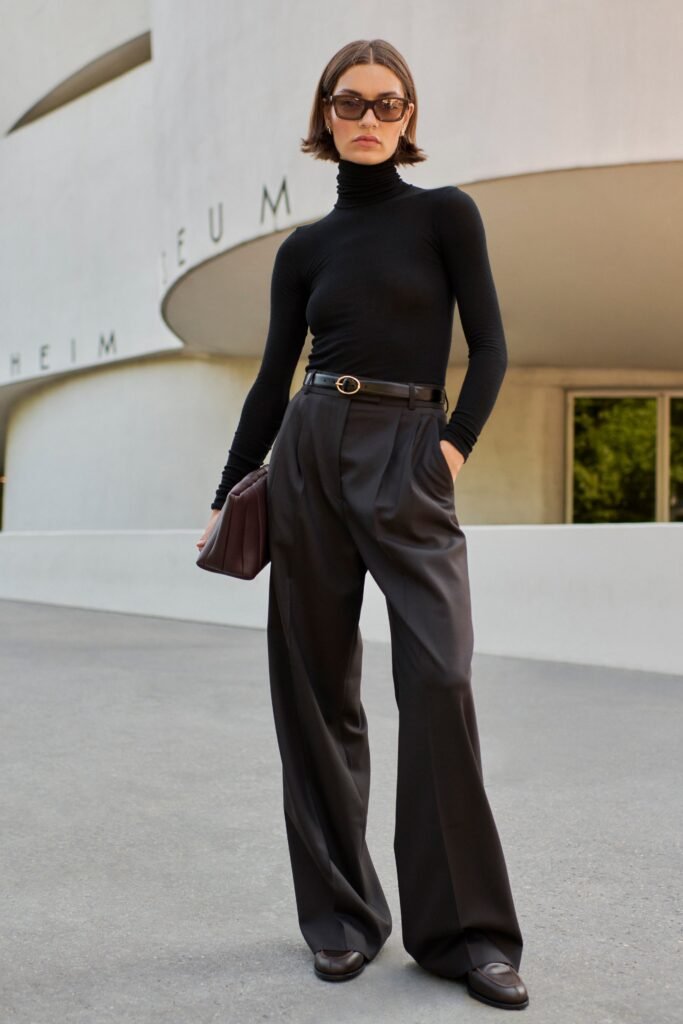 Stylish woman in black turtleneck and wide-leg trousers, posing outdoors in front of modern architecture.