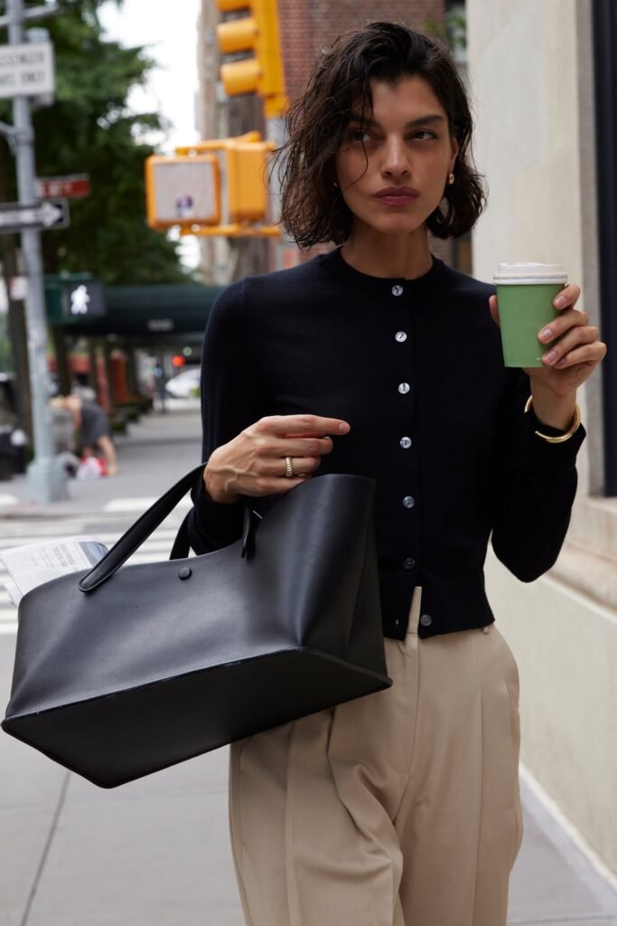 Stylish woman wearing a black cardigan and beige trousers, holding a green coffee cup on a city street.