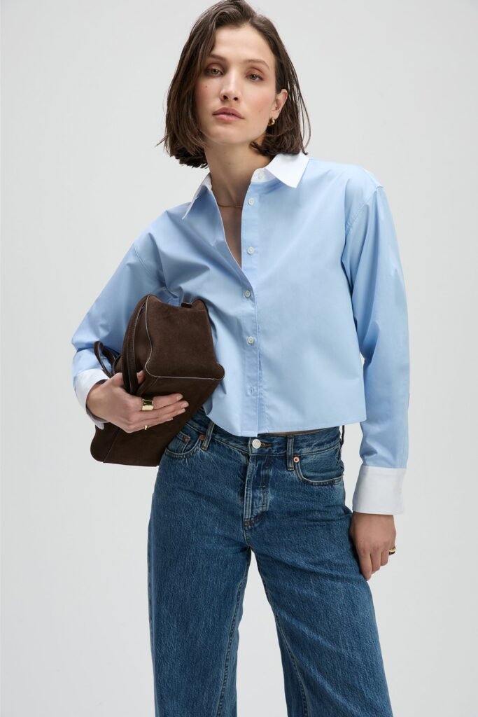 Model in a light blue cropped shirt and denim jeans, holding a brown handbag against a minimalist background.