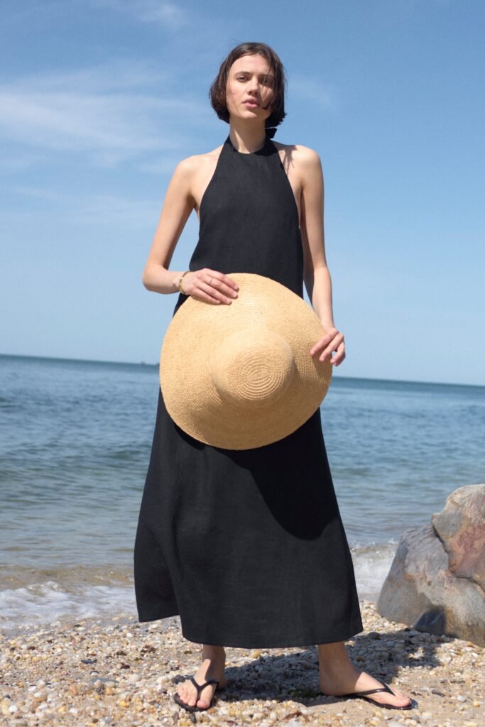 Model in a black halter dress holding a straw hat by the beach, with clear skies and water in the background.