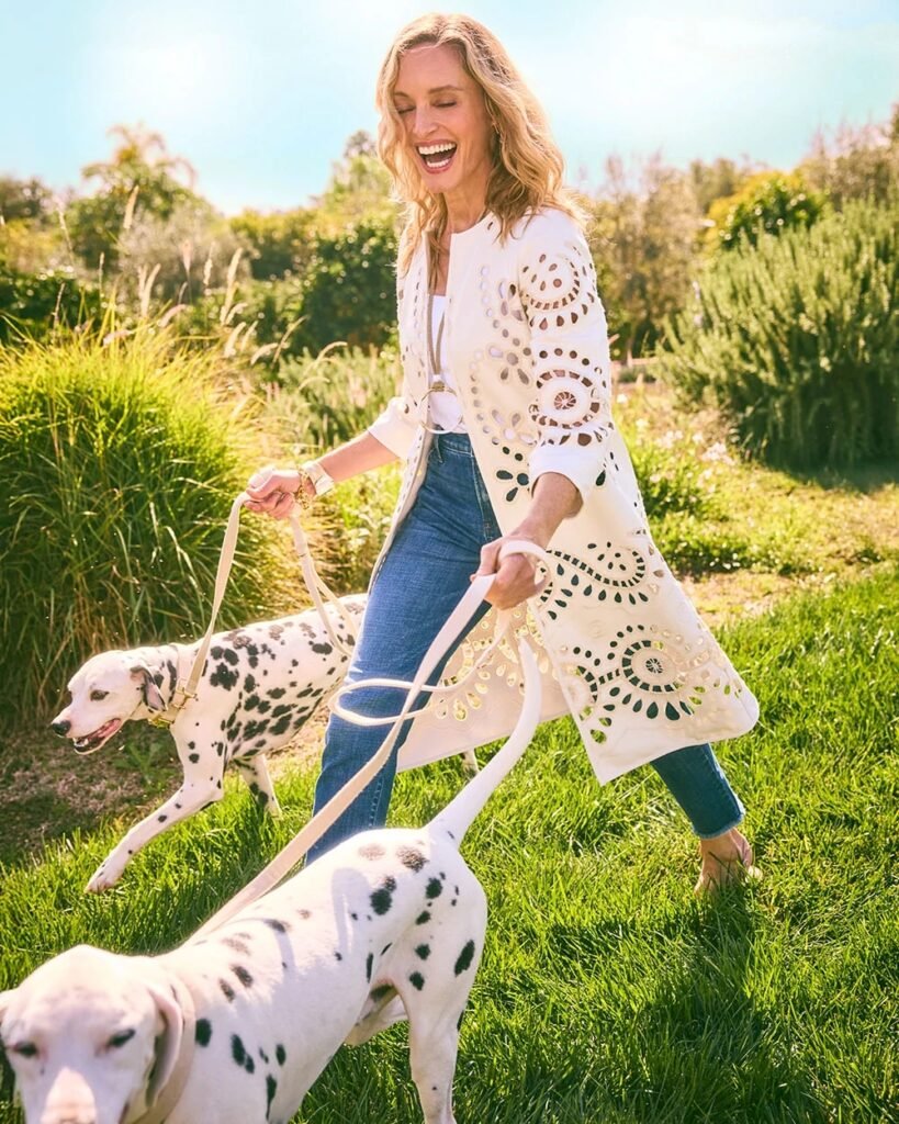 Woman joyfully walking two Dalmatians in a sunny green park, wearing a stylish white jacket and jeans.