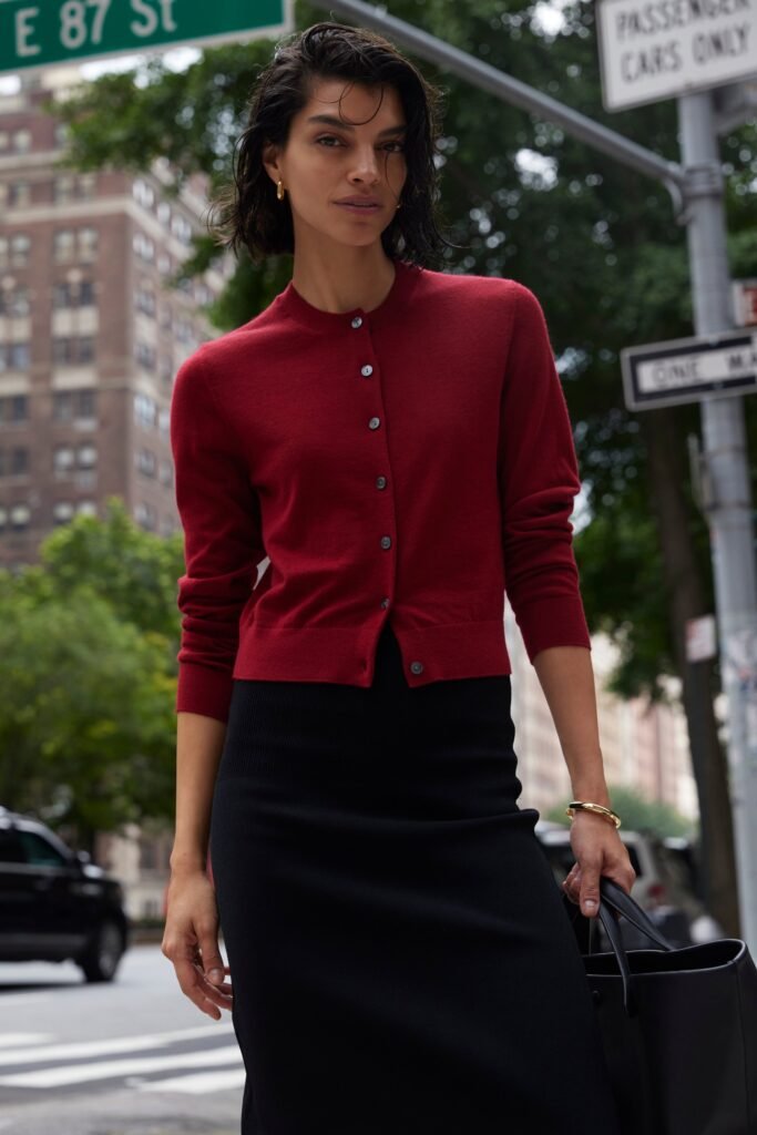 Model wearing a red cardigan and black skirt, posing on a city street with greenery and street signs in the background.